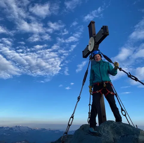 Person standing on a mountain summit under a blue sky with clouds.
