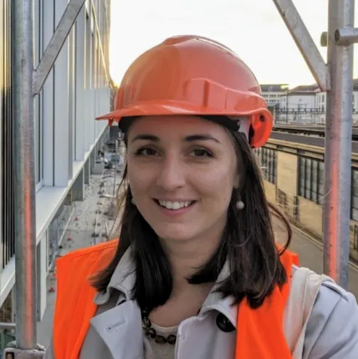 Woman in an orange hard hat and vest standing at a construction site, smiling at the camera.